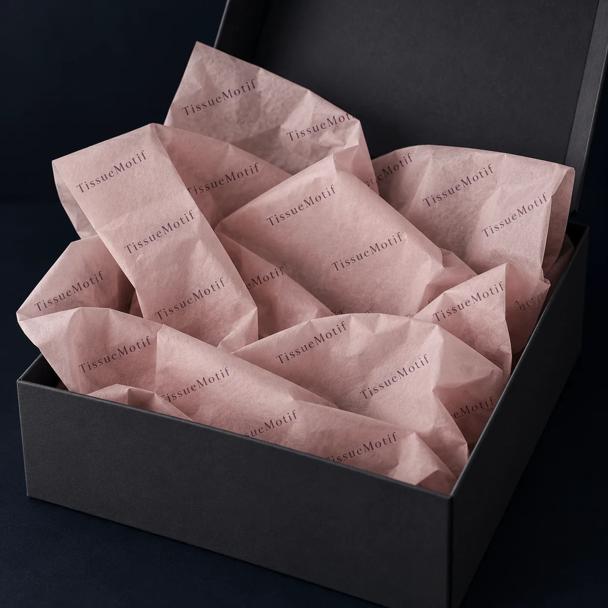 Blush tissue printed with a single dark wordmark, folded inside an open charcoal gift box