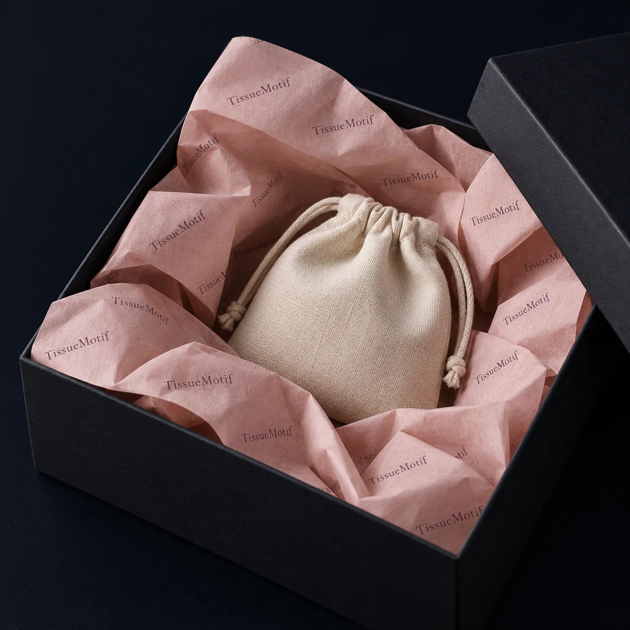 Blush printed tissue folded around a small cream linen pouch in a charcoal gift box